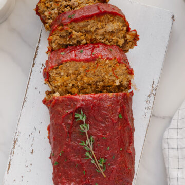 A loaf of vegan meatloaf topped with tomato sauce, partially sliced, is displayed on a white cutting board. A sprig of fresh thyme garnishes the top. The background includes a napkin and marble surface.