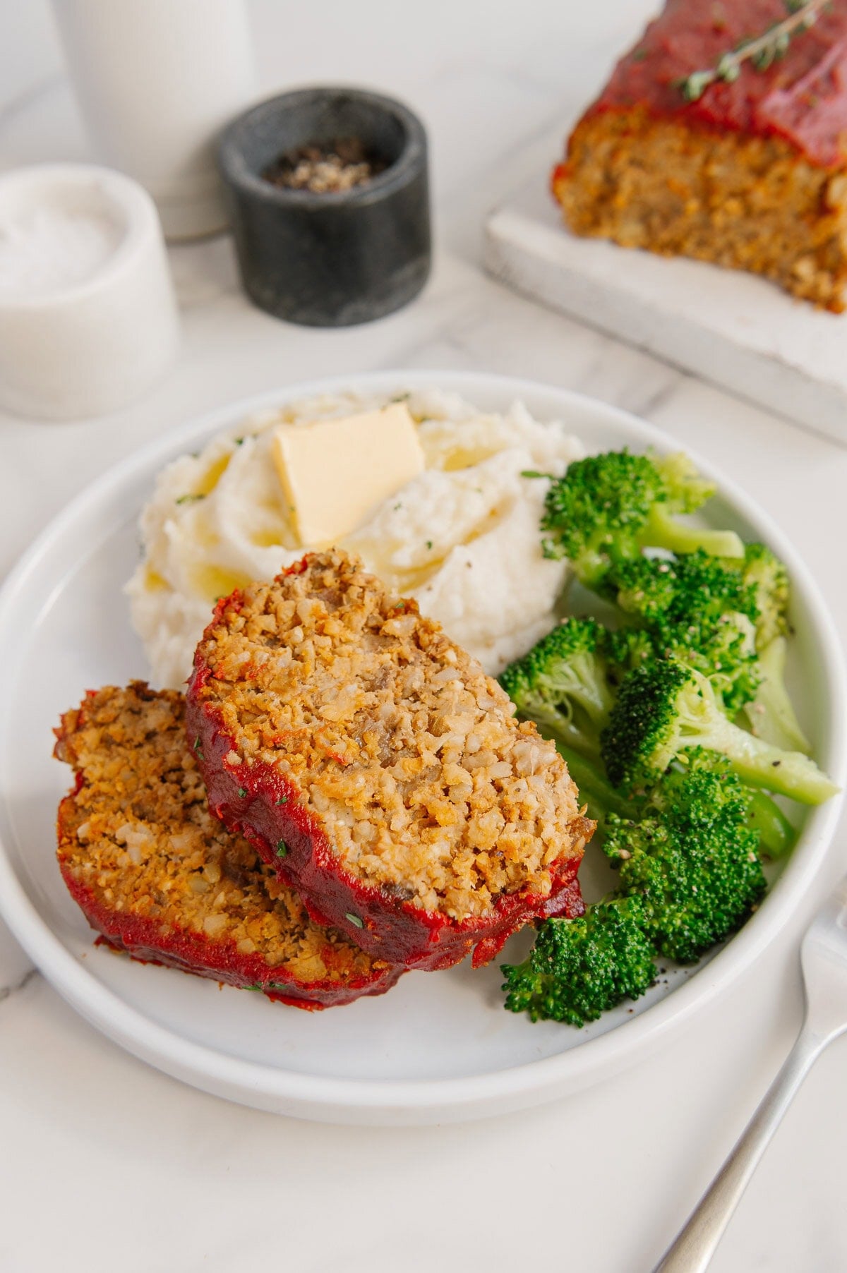 A white plate with two slices of vegan meatloaf, mashed potatoes topped with a pat of butter, and steamed broccoli. In the background are salt, pepper, and more meatloaf on a white board.