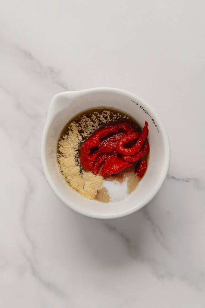 A white bowl on a marble surface contains tomato paste, garlic powder, salt, and maple syrup ready to be mixed.
