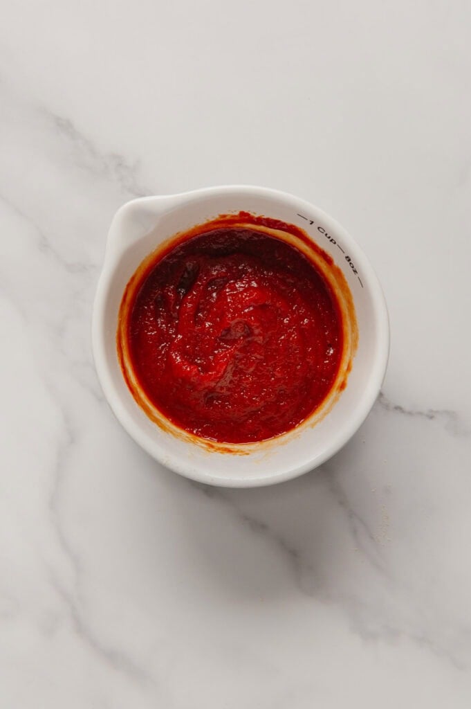 A small white measuring cup filled with thick red glaze on a white marble surface.
