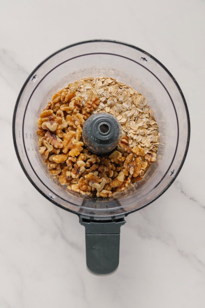 A top-down view of a food processor bowl containing raw walnuts and rolled oats on a white marble surface.