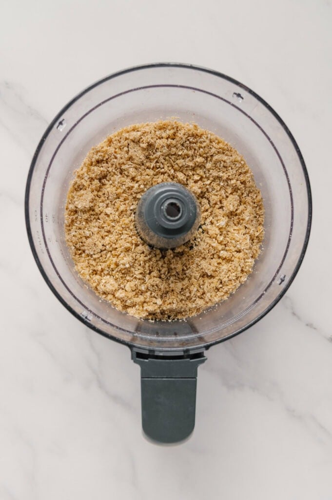 Top view of a food processor bowl containing finely crushed graham crackers or cookie crumbs on a white marble surface.