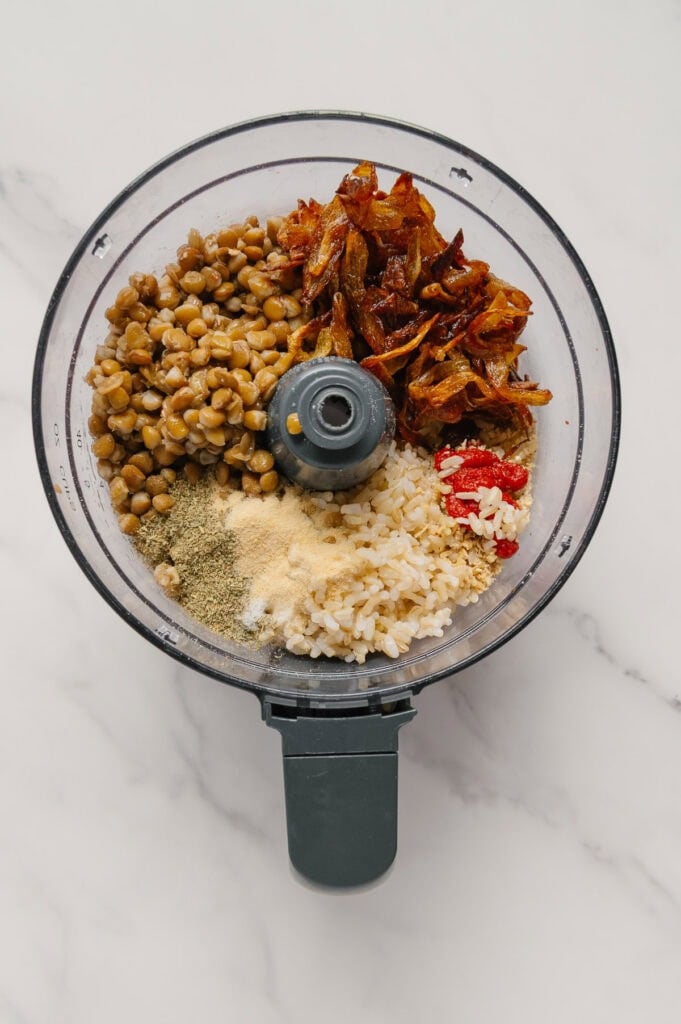 A food processor bowl containing lentils, caramelized onions, cooked rice, tomato paste, garlic powder, and dried herbs, all ready to be blended.
