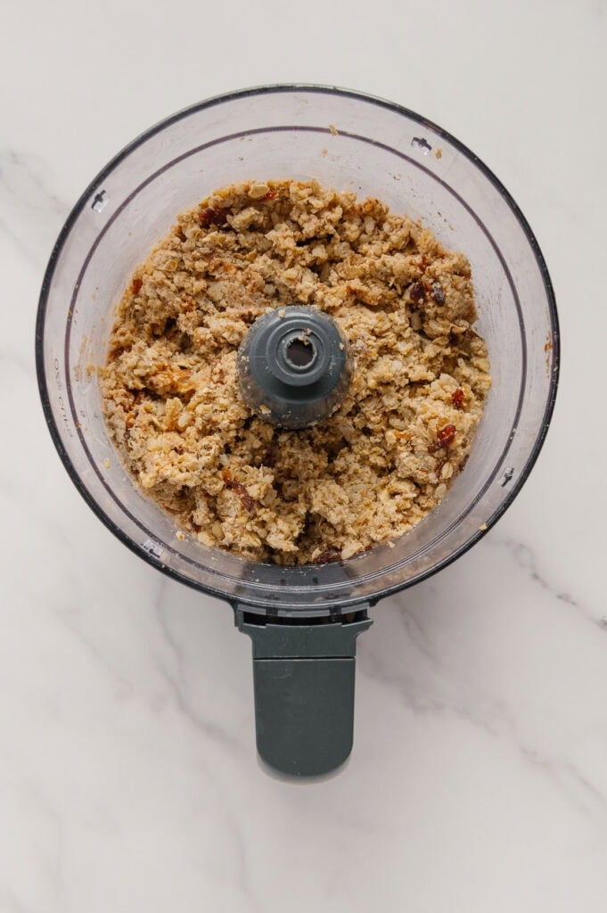 Top view of a food processor containing a chunky, crumbly mixture on a white marble surface.