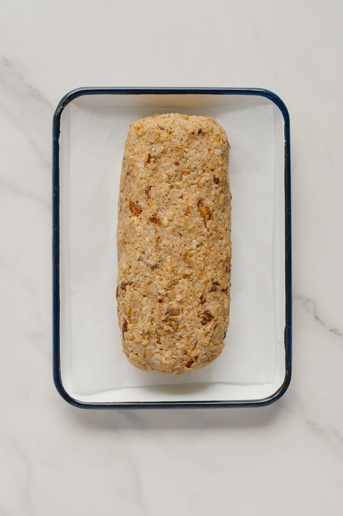 A rectangular loaf of uncooked vegan meatloaf sits on white parchment paper in a black-rimmed baking pan on a marble surface.