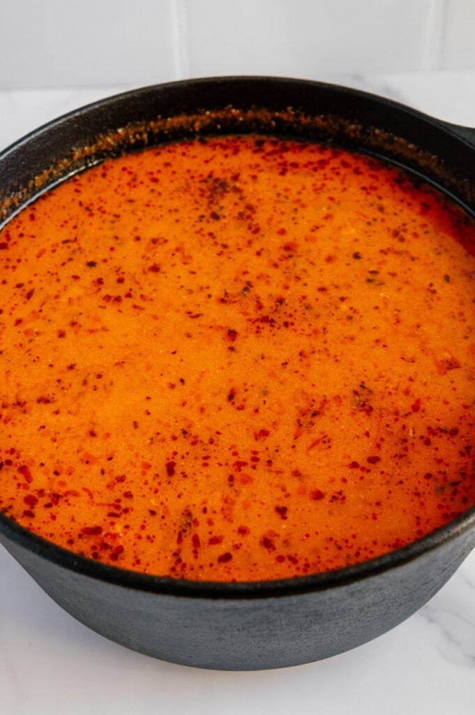 A close-up of a pot filled with bright red-orange, oily, and slightly speckled soup or stew, likely a spicy dish, in a black cast iron pot on a white surface.