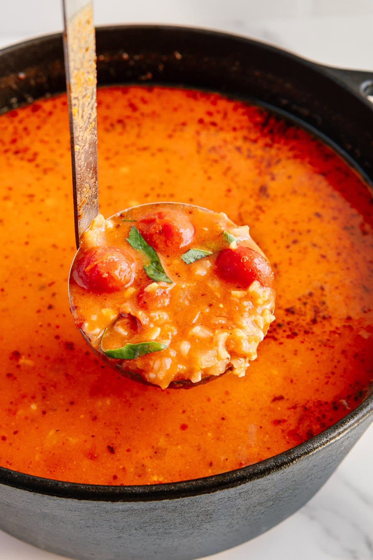 A ladle lifts a serving of creamy vegan tomato soup with rice, cherry tomatoes, and fresh herbs from a black pot filled with orange-red broth.