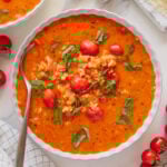 A bowl of creamy vegan tomato soup with rice, cherry tomatoes, and fresh basil, next to a spoon, garlic cloves, vegan parmesan cheese, cherry tomatoes, and a black pepper grinder on a marble surface.