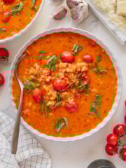 A bowl of creamy vegan tomato soup with rice, cherry tomatoes, and fresh basil, next to a spoon, garlic cloves, vegan parmesan cheese, cherry tomatoes, and a black pepper grinder on a marble surface.