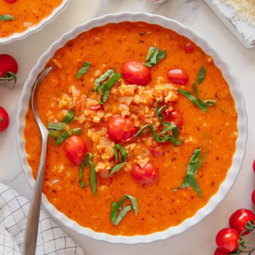 A bowl of creamy vegan tomato soup with rice, cherry tomatoes, and fresh basil, next to a spoon, garlic cloves, vegan parmesan cheese, cherry tomatoes, and a black pepper grinder on a marble surface.
