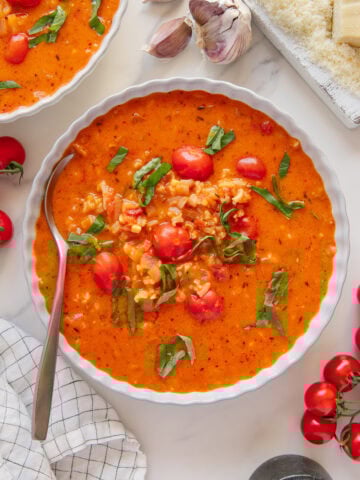 A bowl of creamy vegan tomato soup with rice, cherry tomatoes, and fresh basil, next to a spoon, garlic cloves, vegan parmesan cheese, cherry tomatoes, and a black pepper grinder on a marble surface.