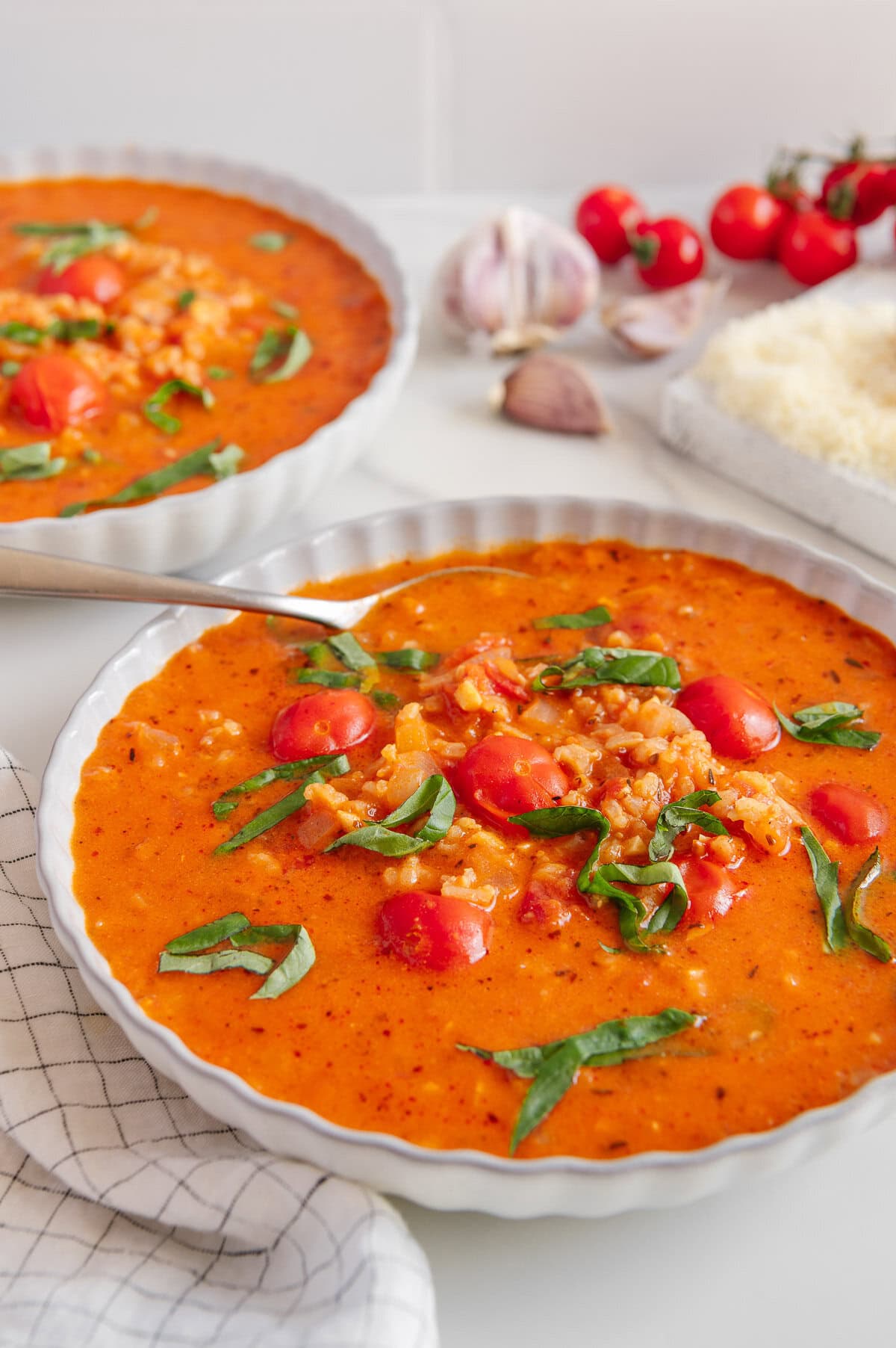 A bowl of creamy vegan tomato soup with cherry tomatoes, rice, and fresh basil leaves, with another bowl in the background. Garlic, tomatoes, and grated vegan cheese are also visible on the table.