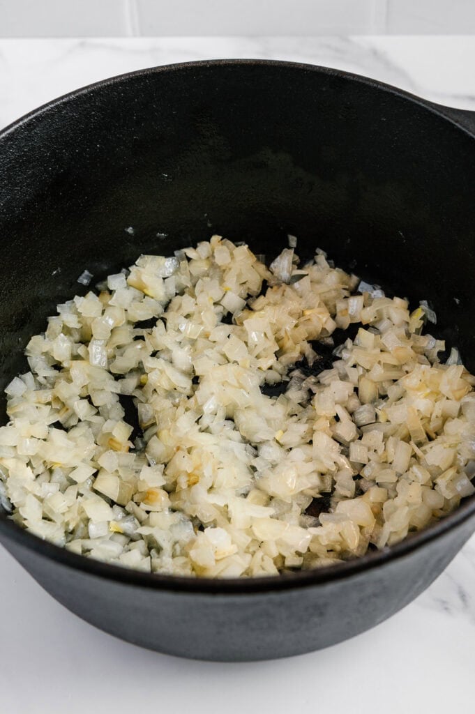 Diced onions being sautéed in a black cast iron pot, with the onions turning translucent on a white countertop.