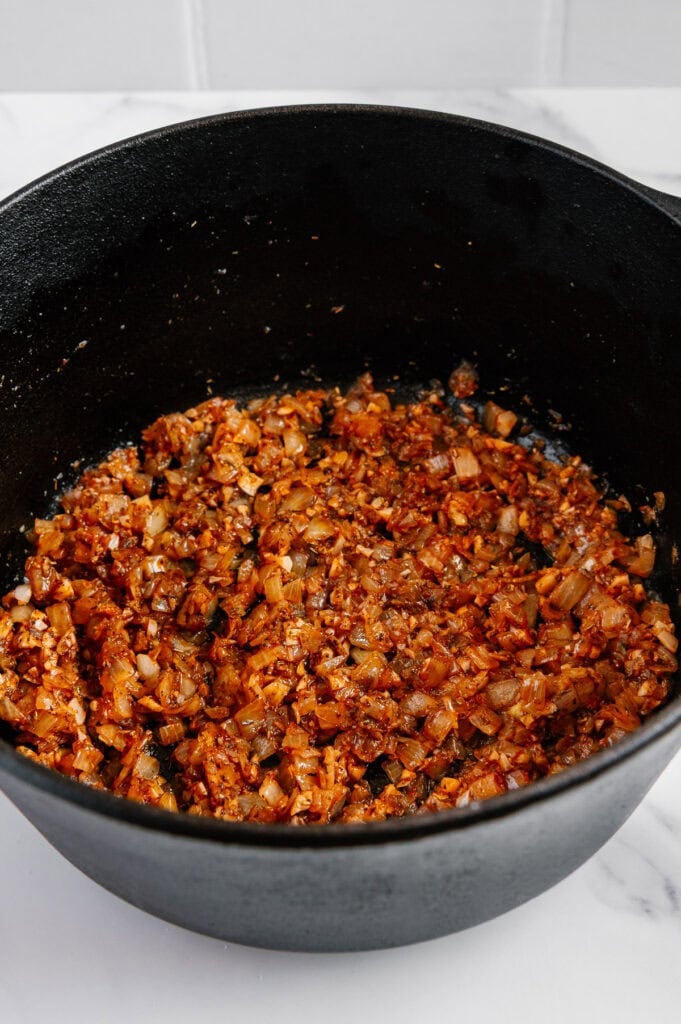 Chopped onions and spices are sautéing in a black pot, creating a golden-brown mixture on a white surface with a tiled background.
