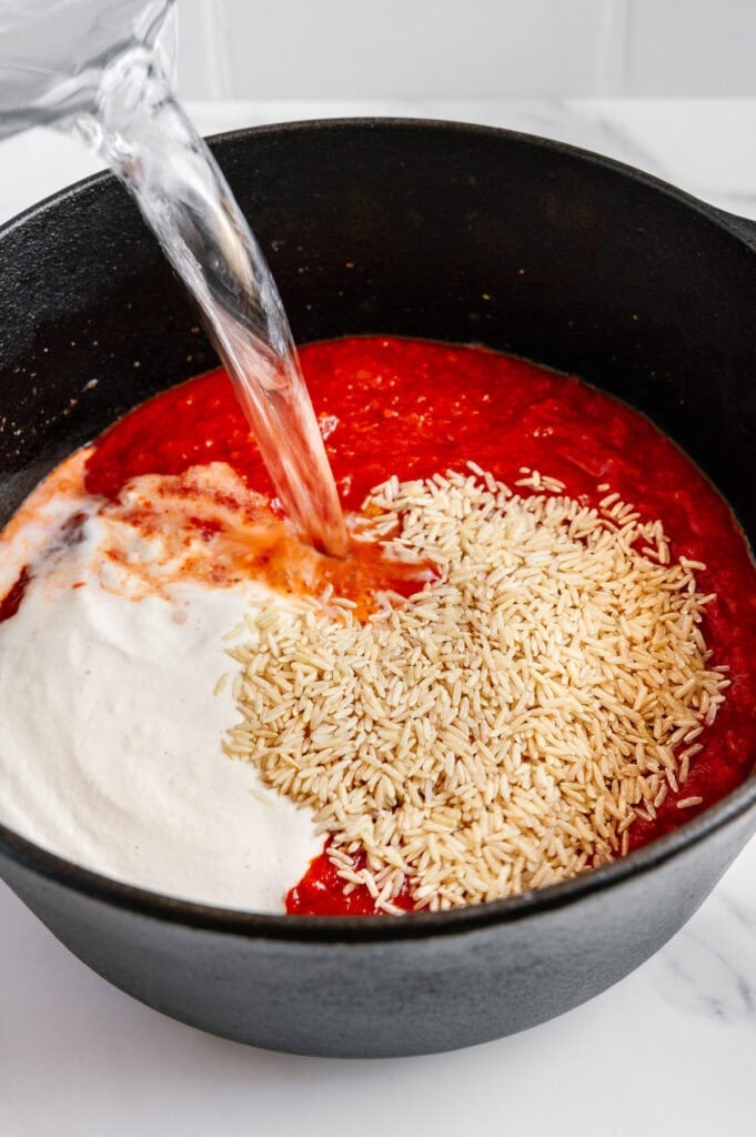 A close-up of a black pot filled with tomato sauce, uncooked rice, and a creamy white sauce, while water is being poured in from the left.
