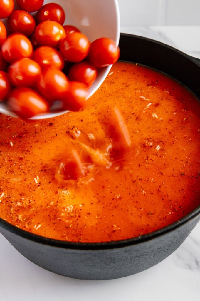 A white bowl of cherry tomatoes is being poured into a black pot filled with orange-red tomato soup or sauce, creating a splash. The scene is set on a white countertop.