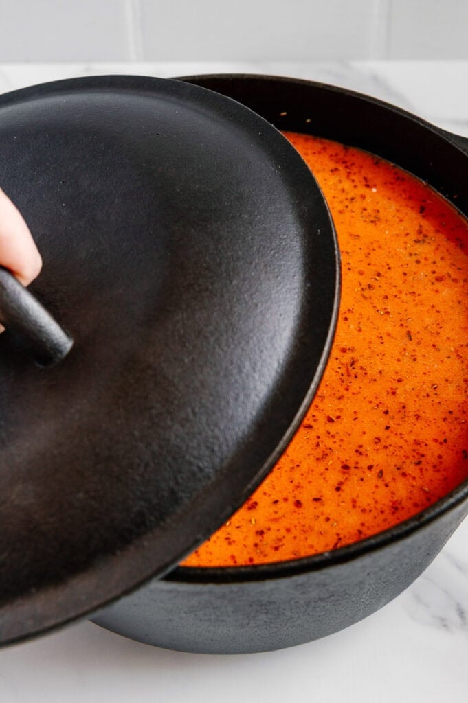 A hand lifts the lid off a black cast iron pot filled with vibrant orange-red soup or sauce, placed on a white marble surface.