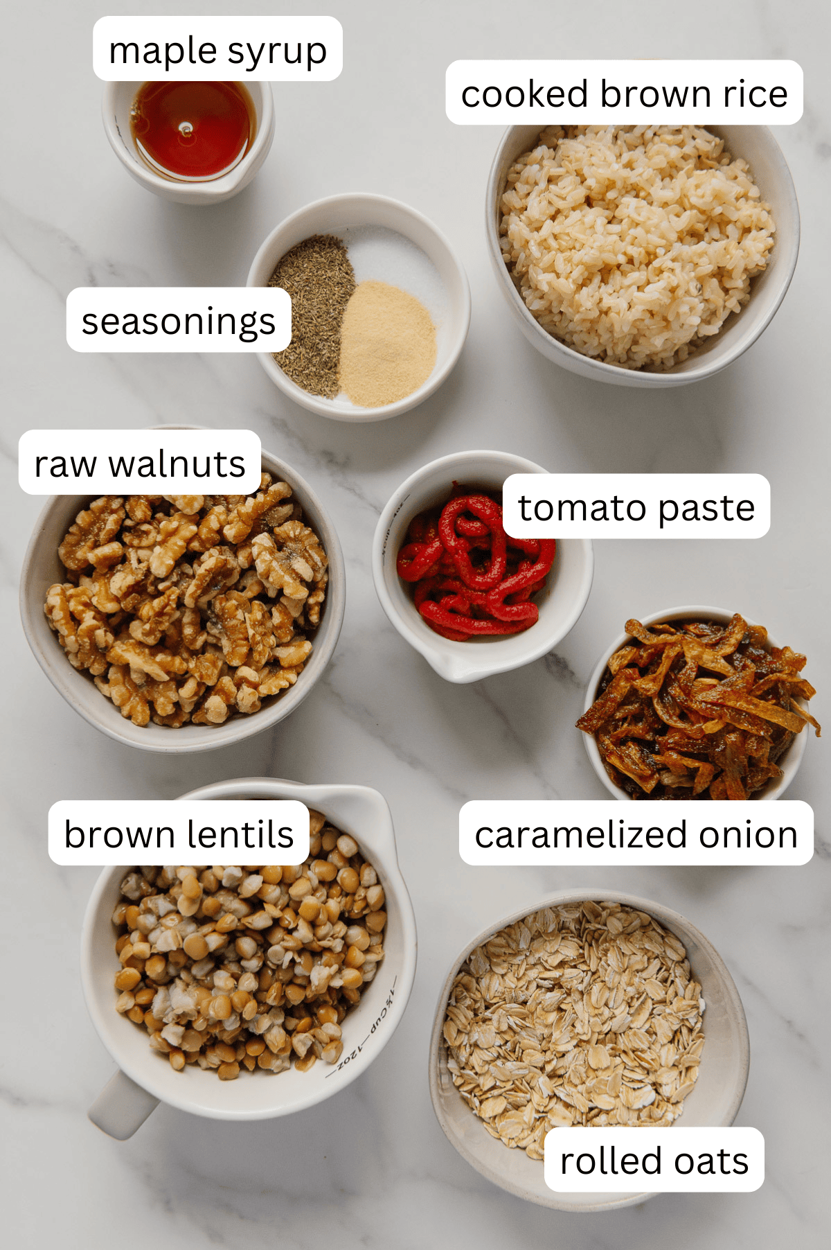 Seven bowls on a white surface contain labeled ingredients for a Vegan Meatloaf: maple syrup, cooked brown rice, seasonings, raw walnuts, tomato paste, brown lentils, caramelized onion, and rolled oats. A hearty vegan recipe in the making!.