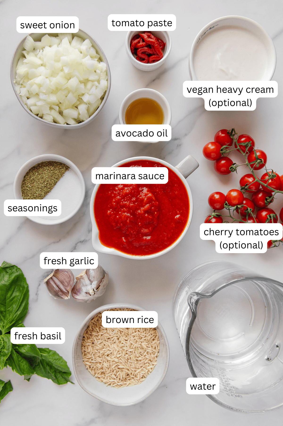 Flat lay of labeled plant-based ingredients for vegan tomato rice soup on a white surface: chopped sweet onion, tomato paste, vegan heavy cream, avocado oil, seasonings, marinara sauce, cherry tomatoes, fresh garlic, fresh basil, brown rice, and water.
