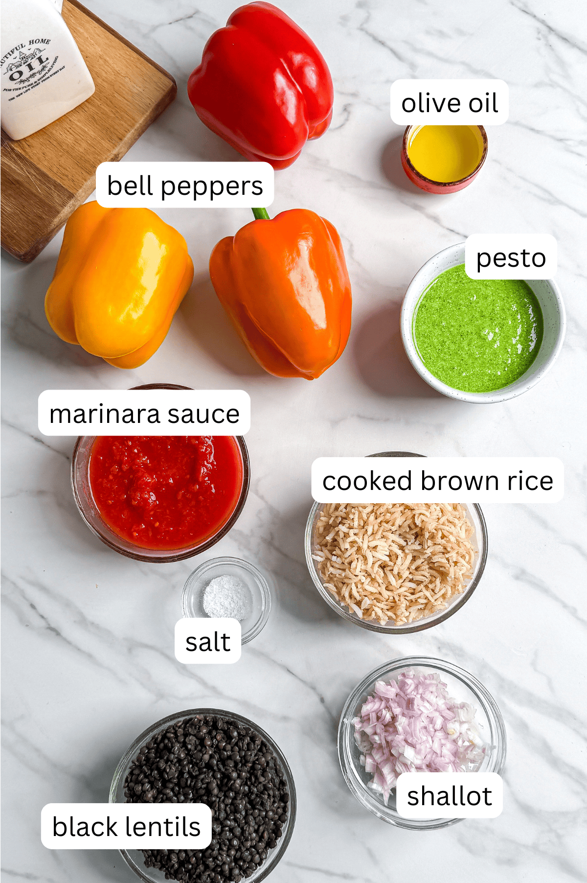Top-down view of ingredients for Italian Lentil Stuffed Bell Peppers on a marble surface-red, yellow, and orange bell peppers, olive oil, pesto, marinara sauce, brown rice, salt, black lentils, and chopped shallot. Each item is labeled.