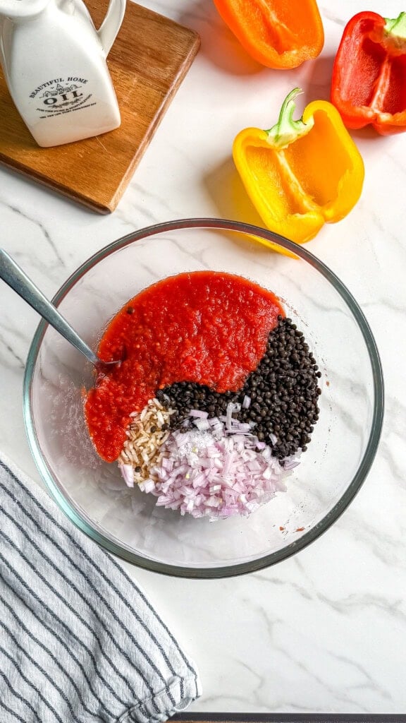 A glass bowl with tomato sauce, black lentils, chopped onions, and brown rice being mixed with a spoon on a marble counter, next to halved red and yellow bell peppers and a striped towel.