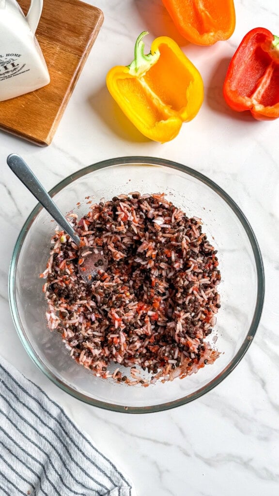 A glass bowl filled with a mixture of rice and black beans sits on a marble countertop, with a spoon inside. Nearby are a striped towel, a wooden cutting board, and halved yellow and red bell peppers.