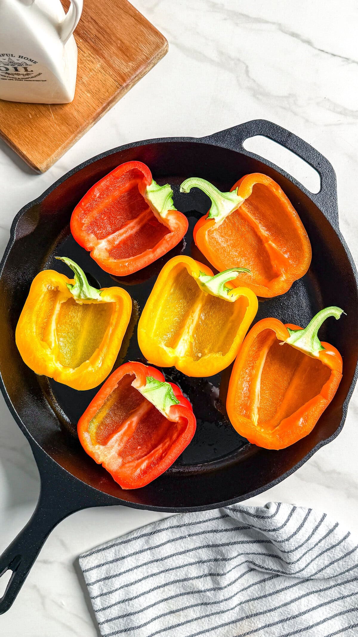 Halved red, orange, and yellow bell peppers arranged in a black cast iron skillet on a white surface, with a striped cloth nearby and a wooden cutting board partially visible.