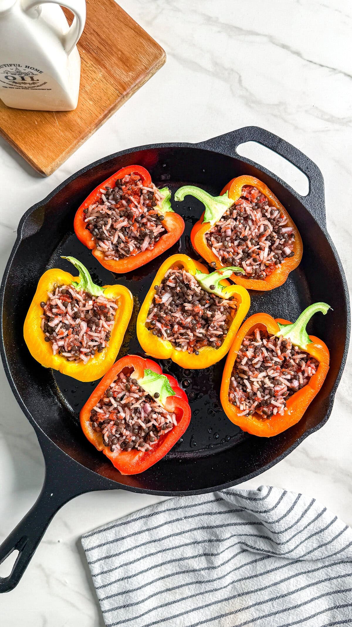 Six bell pepper halves, red and yellow, filled with a mixture of rice and beans, arranged in a black cast iron skillet on a white marble surface with a striped towel and a ceramic container nearby.