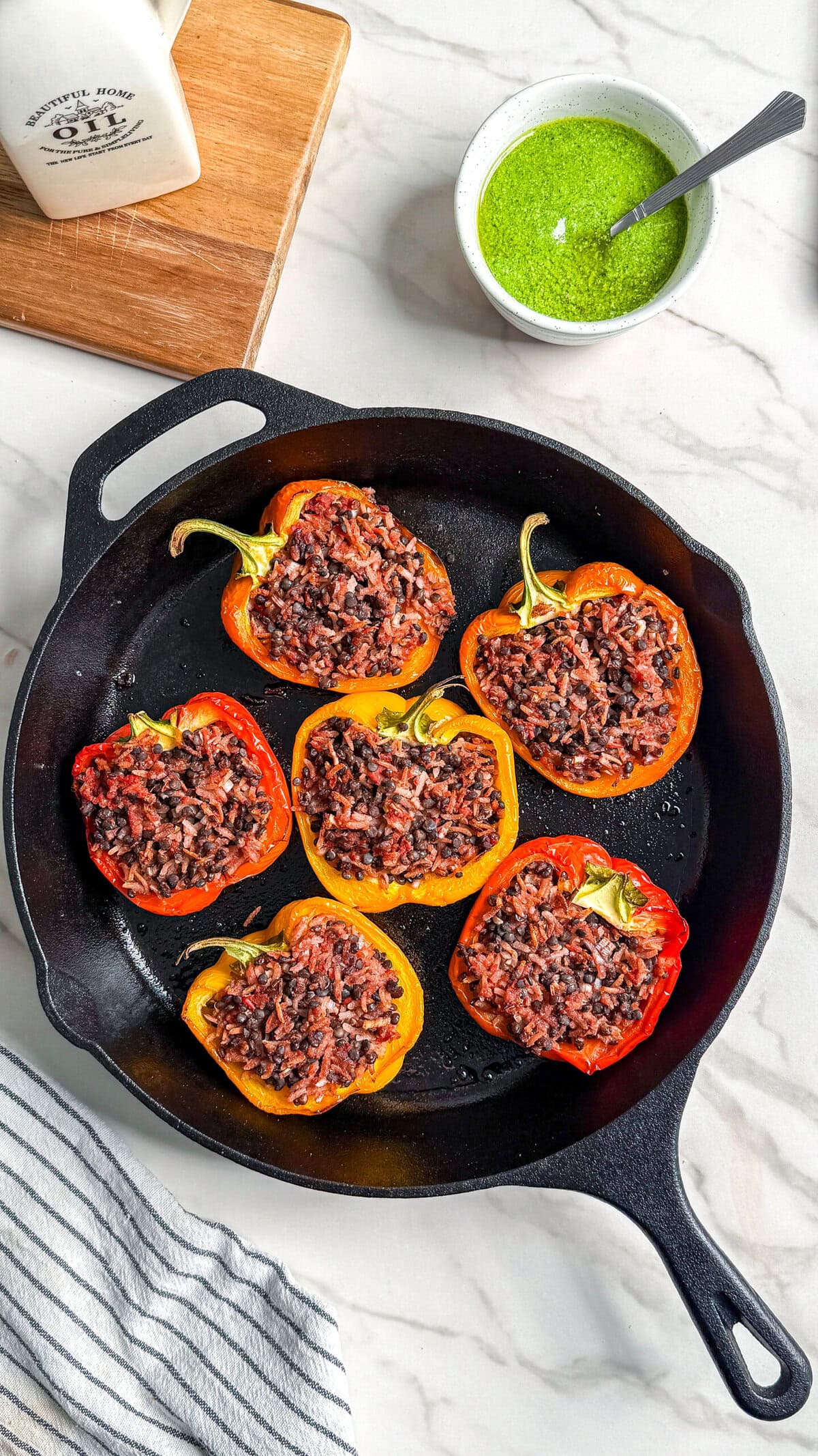 A black cast iron skillet with six halved bell peppers stuffed with a dark rice mixture, sitting on a marble counter. A small bowl of green sauce and a striped towel are nearby.