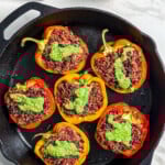 Seven halved bell peppers stuffed with rice and ground meat, topped with green sauce, are arranged in a cast iron skillet. A bowl of green sauce, a wedge of cheese, and a striped towel are nearby on a marble surface.
