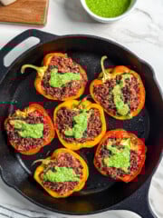 Seven halved bell peppers stuffed with rice and ground meat, topped with green sauce, are arranged in a cast iron skillet. A bowl of green sauce, a wedge of cheese, and a striped towel are nearby on a marble surface.