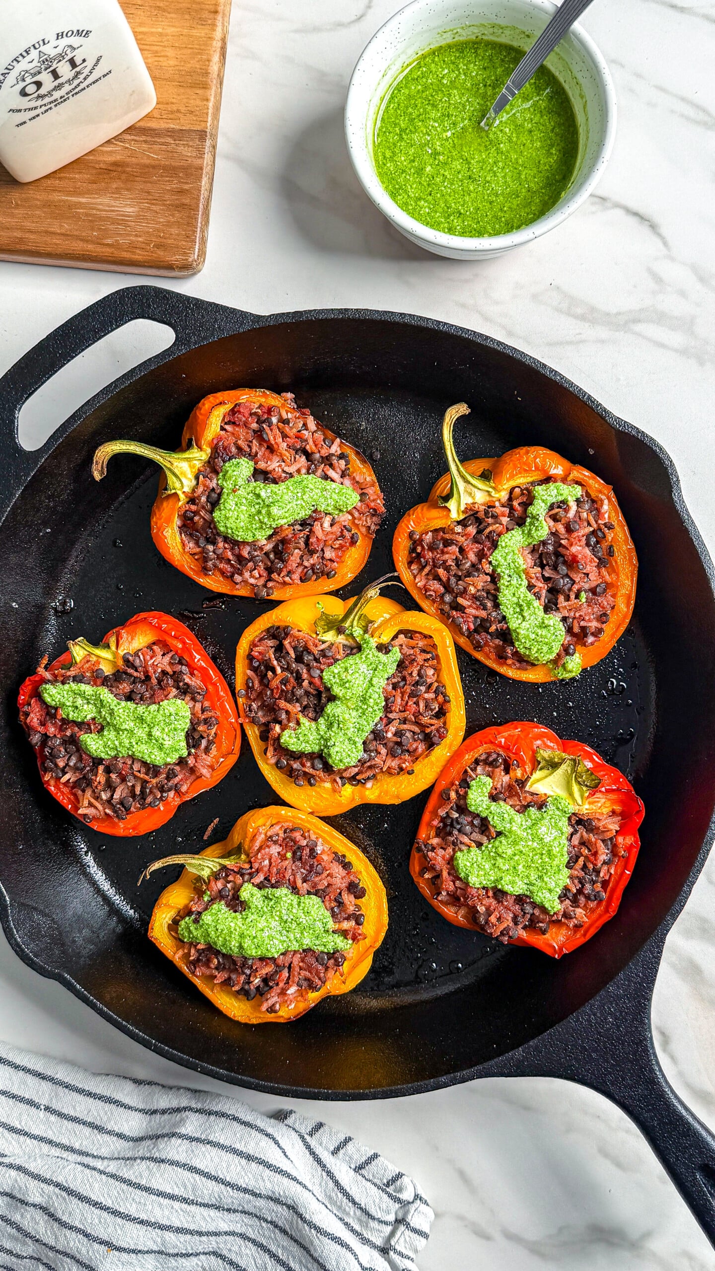 Seven halved bell peppers stuffed with rice and ground meat, topped with green sauce, are arranged in a cast iron skillet. A bowl of green sauce, a wedge of cheese, and a striped towel are nearby on a marble surface.