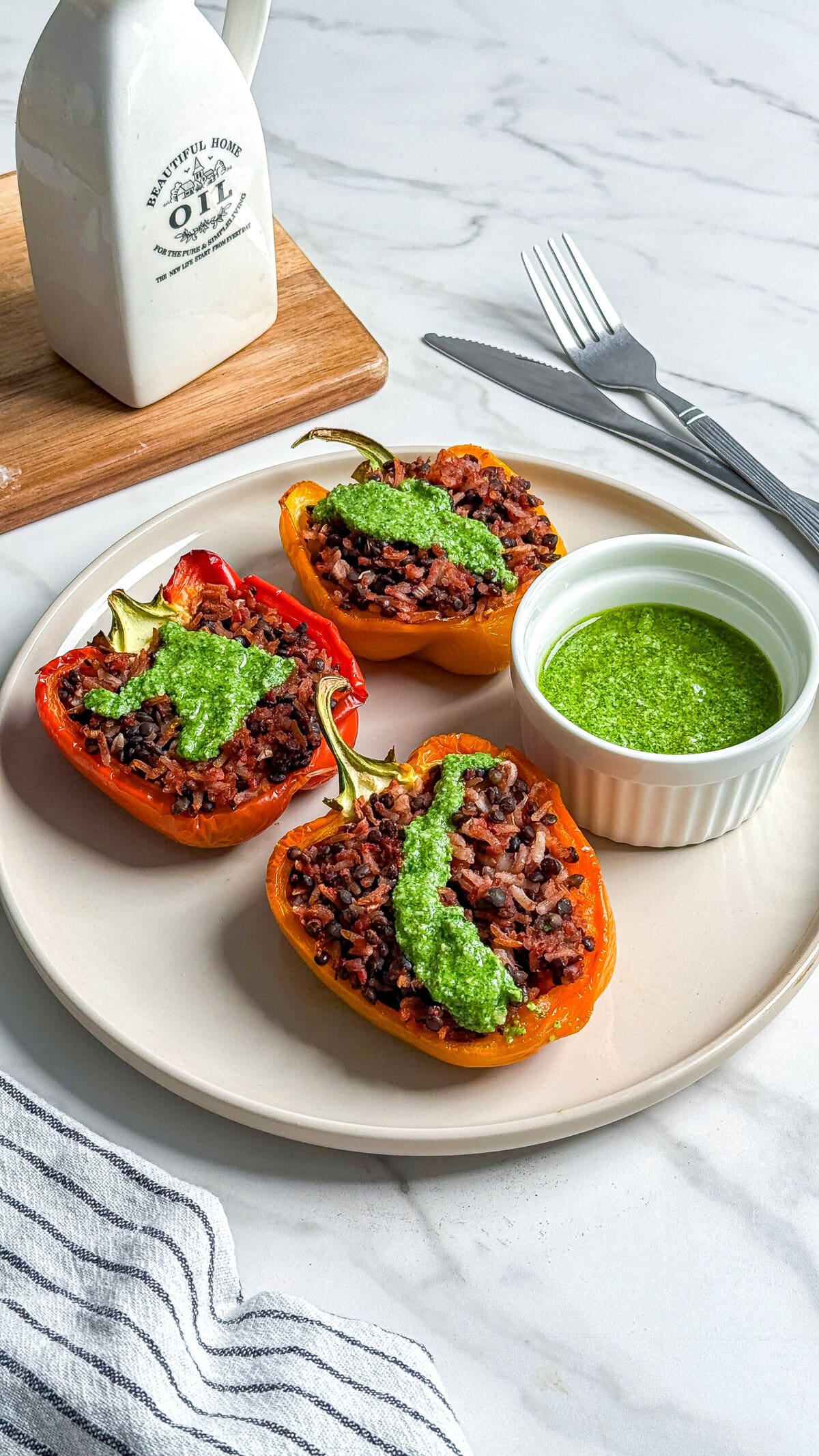 Three halved bell peppers stuffed with rice and topped with green sauce are served on a plate next to a small bowl of green sauce. A fork, knife, striped napkin, and an oil bottle are in the background.