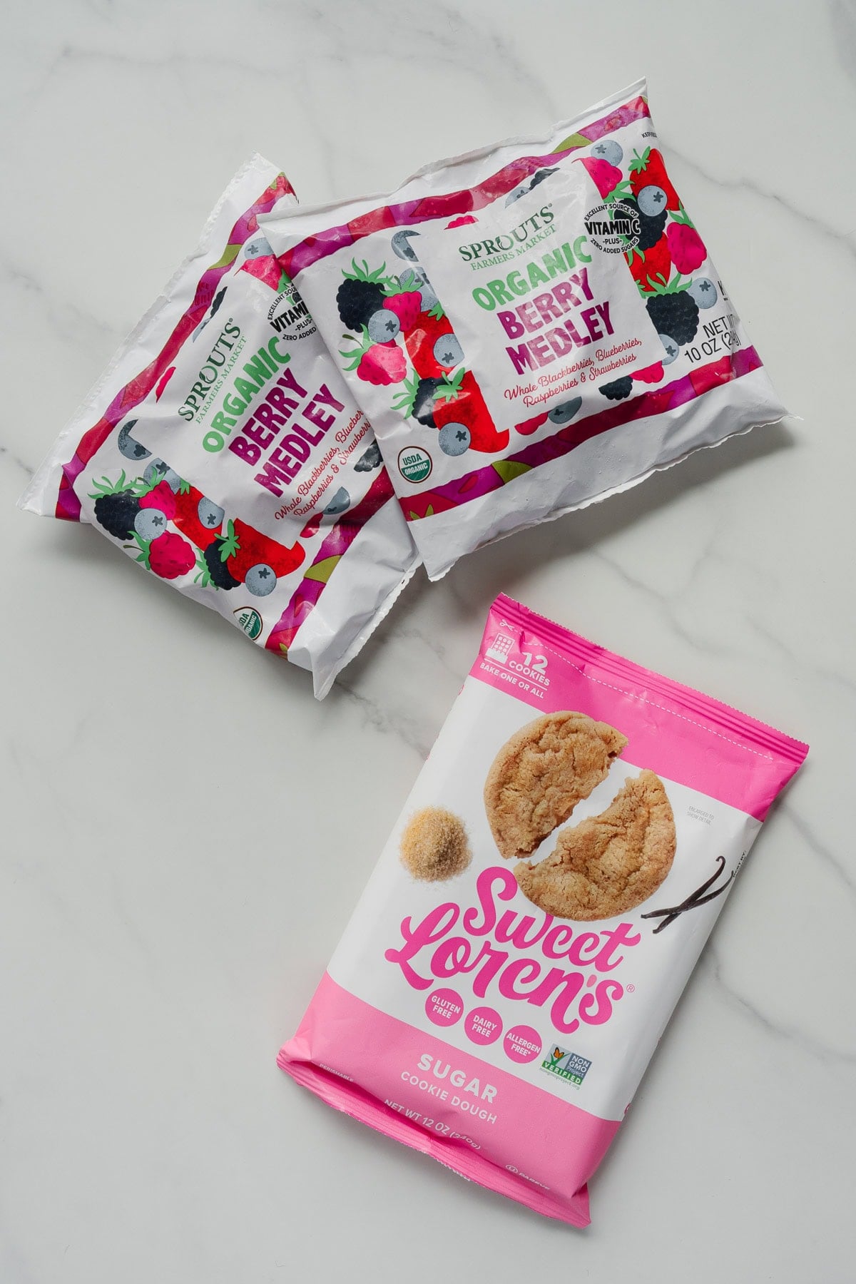 Three grocery items on a white surface: two bags of Sprouts Organic Berry Medley and one pink bag of Sweet Loren's Sugar Cookie Dough, perfect for making a quick 2-ingredient berry cobbler inspired by trending TikTok recipes.