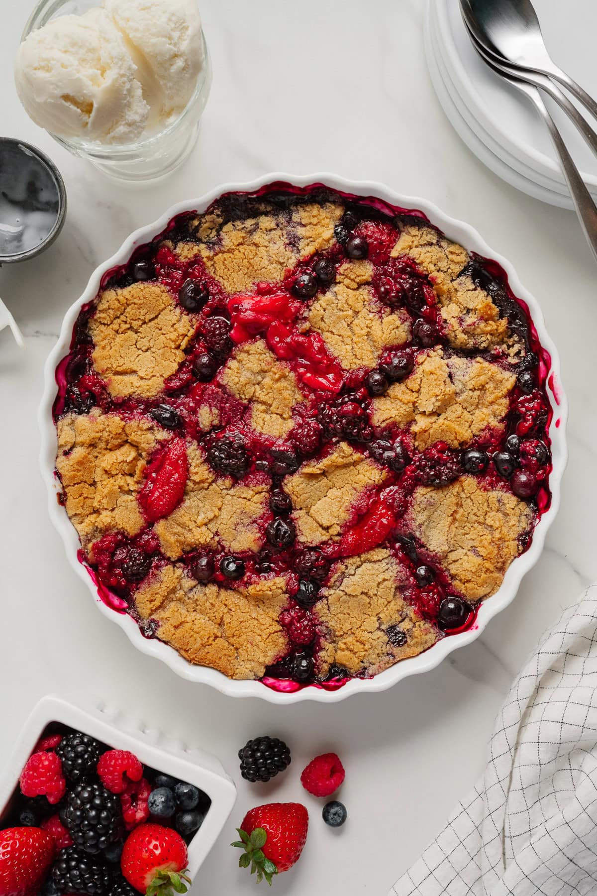 A baked berry cobbler in a white pie dish, featuring golden-brown crust and vibrant berries. Surrounding it are a bowl of vanilla ice cream, fresh berries, a checkered napkin, and stacked plates with spoons—perfect for that viral TikTok recipe moment.