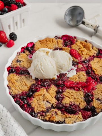 A freshly baked 2-Ingredient Berry Cobbler topped with two scoops of vanilla ice cream in a white pie dish, with a basket of fresh berries and an ice cream scooper in the background on a marble surface-a trending TikTok recipe.