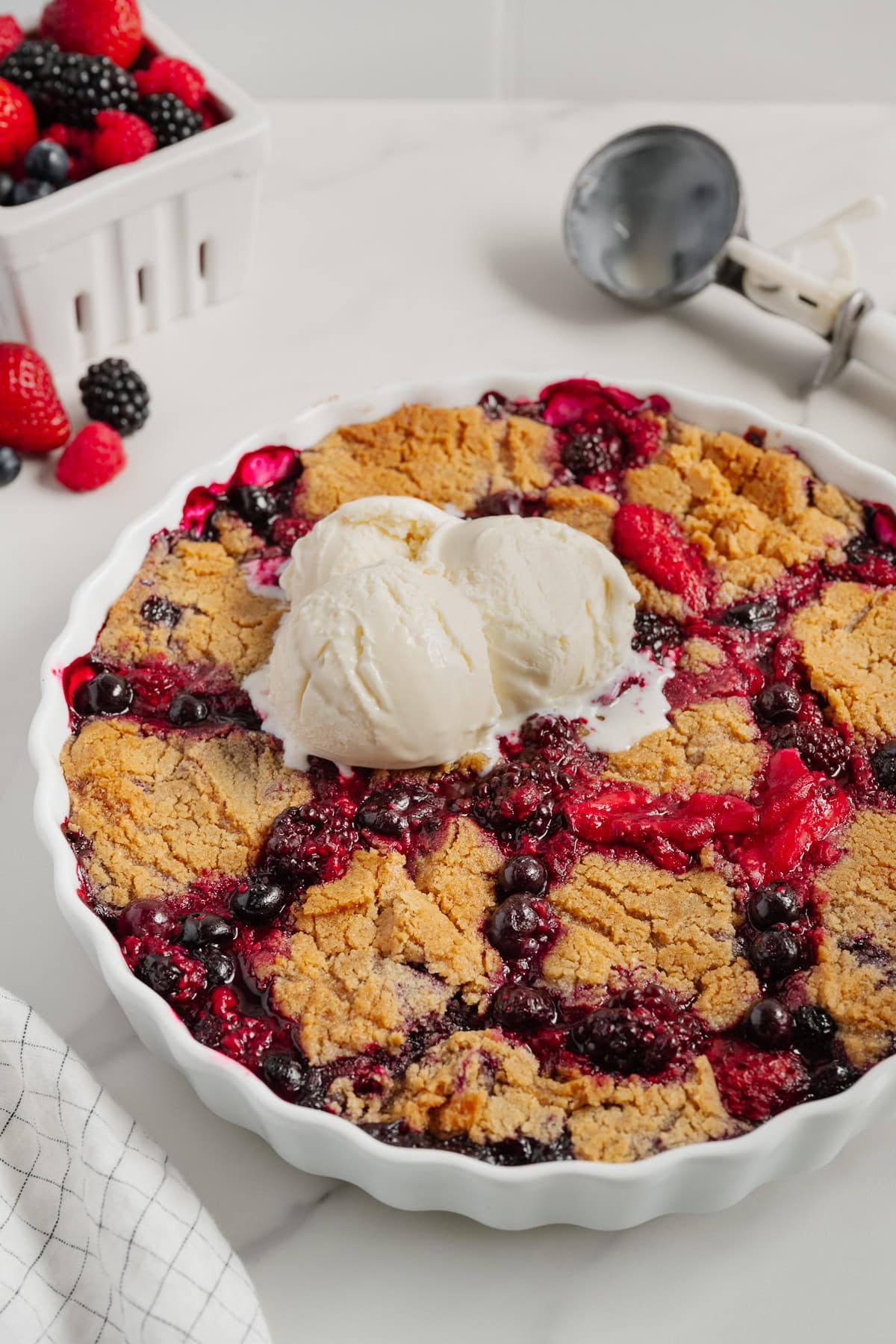 A freshly baked 2-Ingredient Berry Cobbler topped with two scoops of vanilla ice cream in a white pie dish, with a basket of fresh berries and an ice cream scooper in the background on a marble surface—a trending TikTok recipe.