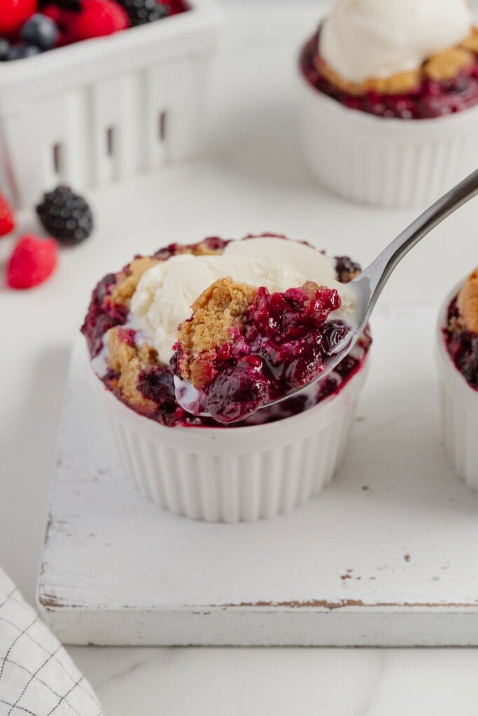 A spoon lifts a bite of berry cobbler topped with vanilla ice cream from a white ramekin. Fresh berries and another serving are visible in the background on a white surface, showcasing this easy TikTok recipe.