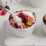 A spoonful of 2-Ingredient Berry Cobbler topped with vanilla ice cream is lifted from a white ramekin, with more cobbler and berries visible in the background on a white surface-an easy TikTok Recipe to try.