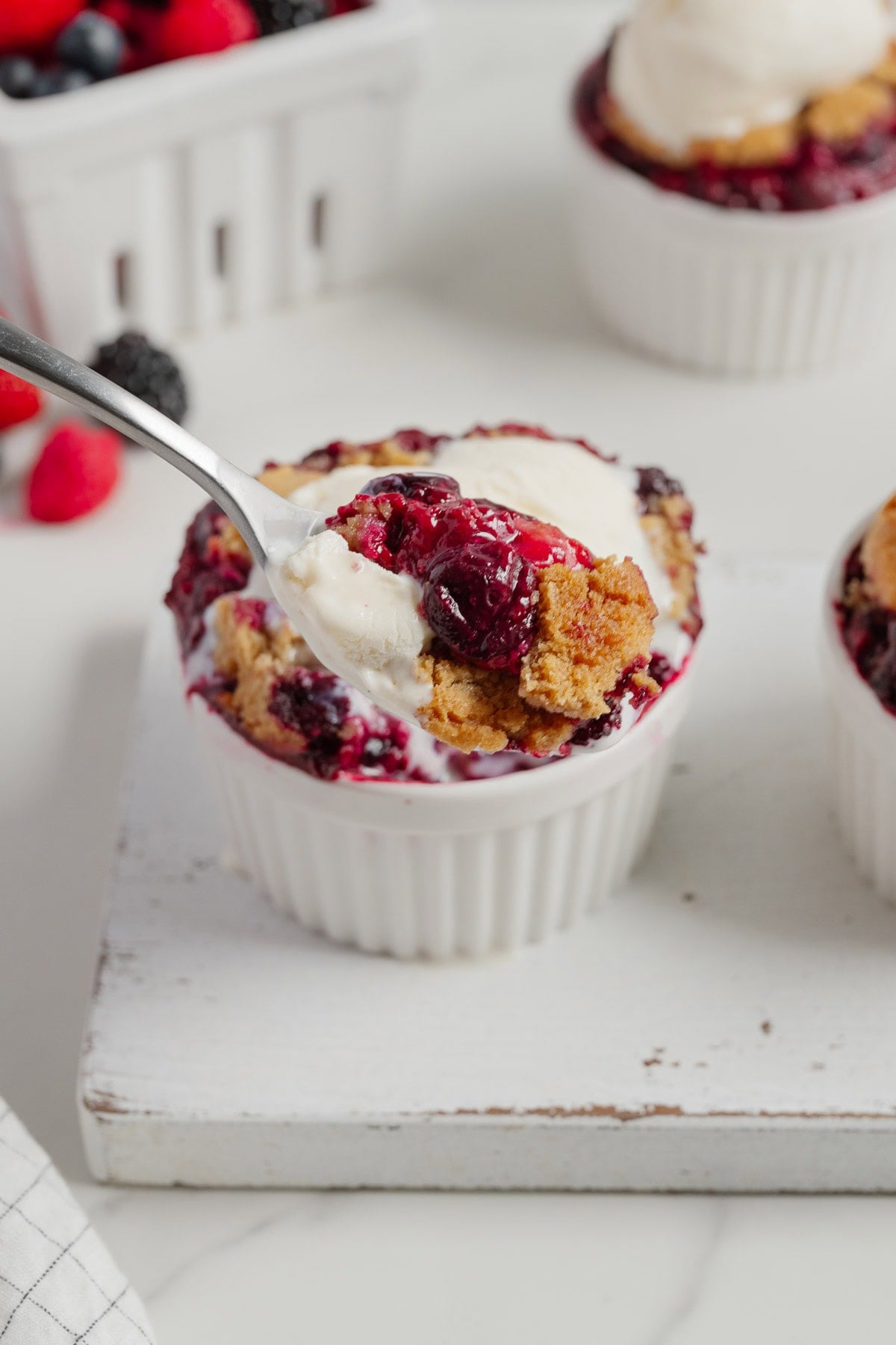 A spoonful of 2-Ingredient Berry Cobbler topped with vanilla ice cream is lifted from a white ramekin, with more cobbler and berries visible in the background on a white surface—an easy TikTok Recipe to try.
