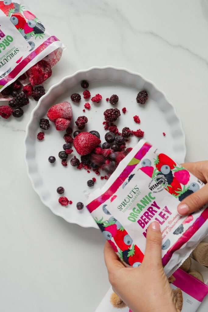 A person's hands pour frozen organic mixed berries from a colorful Sprouts-branded bag into a white pie dish on a marble surface-perfect for a quick 2-Ingredient Berry Cobbler, inspired by the latest TikTok Recipe trend. More berries and an open bag are on the side.