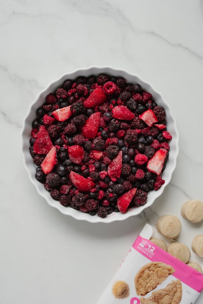 A white tart dish filled with frozen berries sits on a white marble surface, ready for a 2-Ingredient TikTok Recipe. Nearby are round cookies and part of a pink and white cookie package.