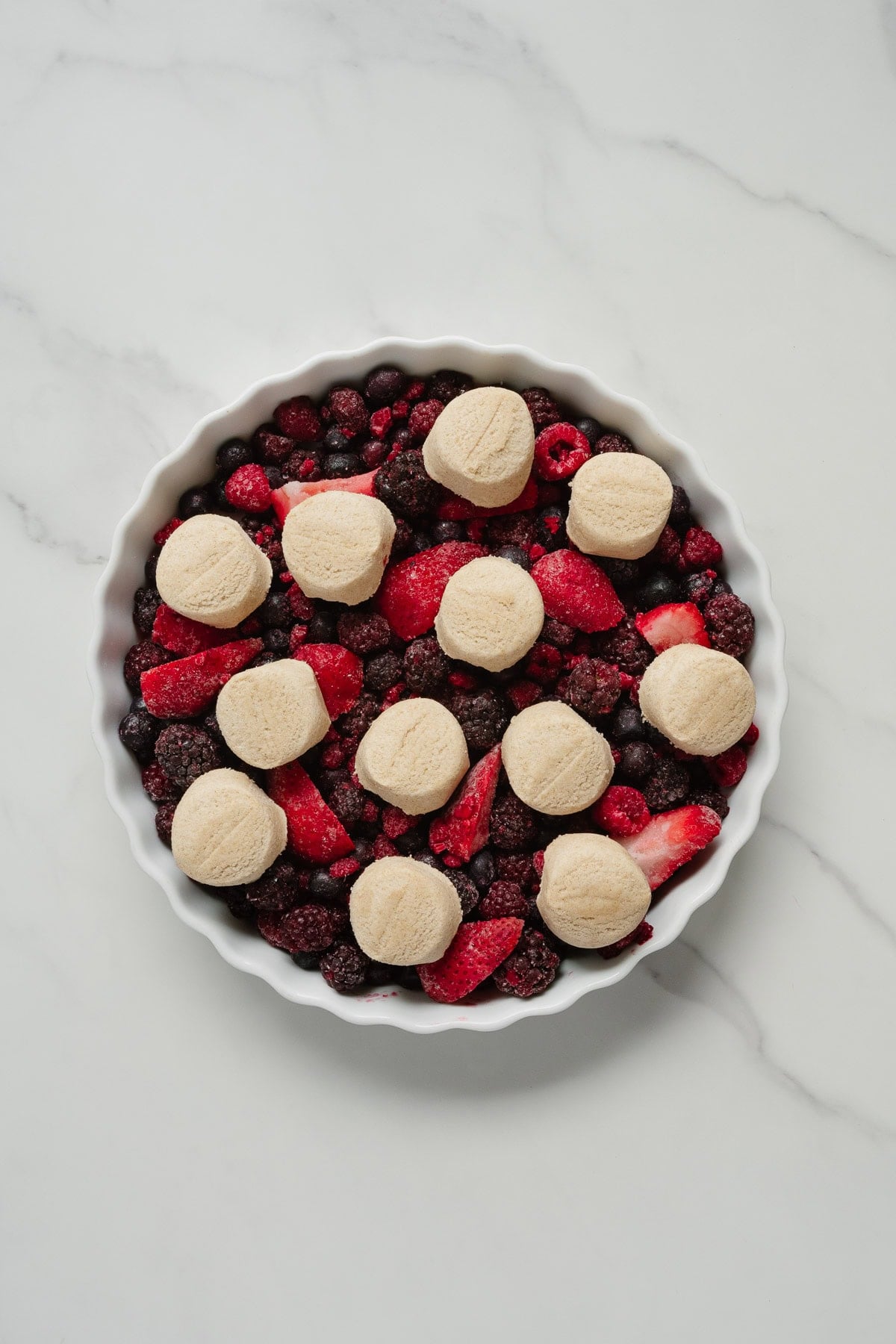 A round white dish filled with mixed berries and topped with evenly spaced dollops of biscuit dough, ready to be baked into a delicious 2-ingredient berry cobbler, on a light marble surface.
