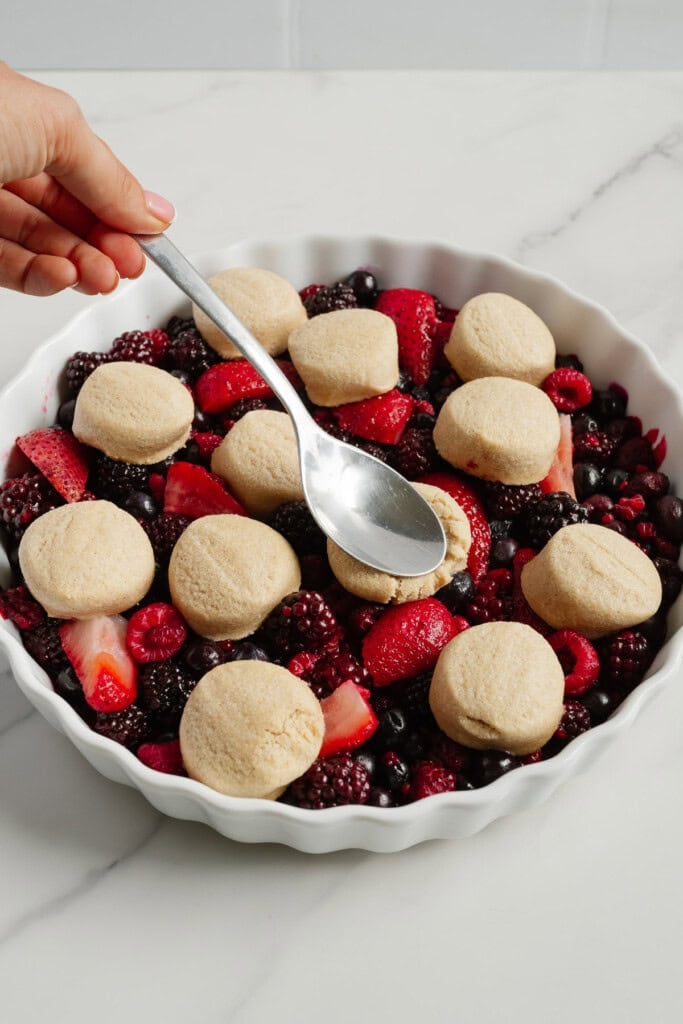 A hand holds a spoon over a round baking dish filled with mixed berries and topped with unbaked biscuit dough, ready to be spread and baked as a delicious 2-Ingredient Berry Cobbler perfect for your next TikTok recipe try.