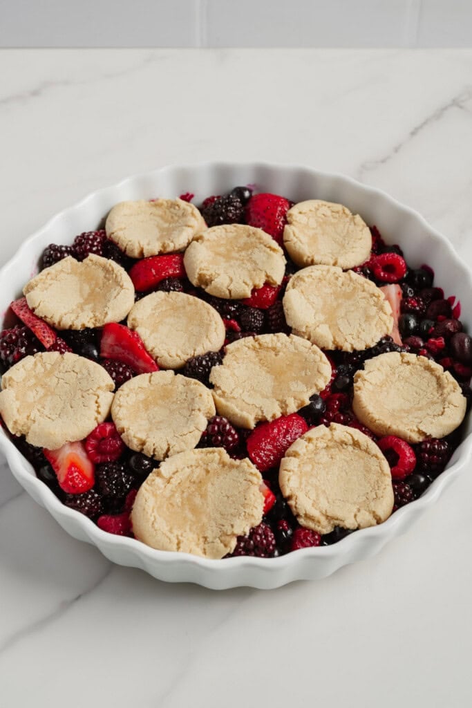 A white baking dish filled with mixed berries and topped with round, unbaked biscuit dough pieces sits on a marble countertop-a classic Berry Cobbler that's trending as an easy TikTok Recipe.