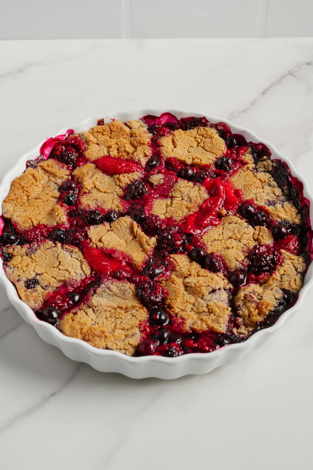 A round baking dish filled with a 2-Ingredient Berry Cobbler, featuring golden-brown biscuit topping and bubbling, vibrant red and purple berry filling on a white marble surface-a trending TikTok recipe everyone loves.