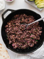 A cast iron skillet filled with cooked, mashed black beans and diced onions, with a spoon resting inside. Nearby are sliced avocado, diced red onion, sour cream, a tortilla, and a checkered towel.
