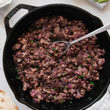 A cast iron skillet filled with cooked, mashed black beans and diced onions, with a spoon resting inside. Nearby are sliced avocado, diced red onion, sour cream, a tortilla, and a checkered towel.