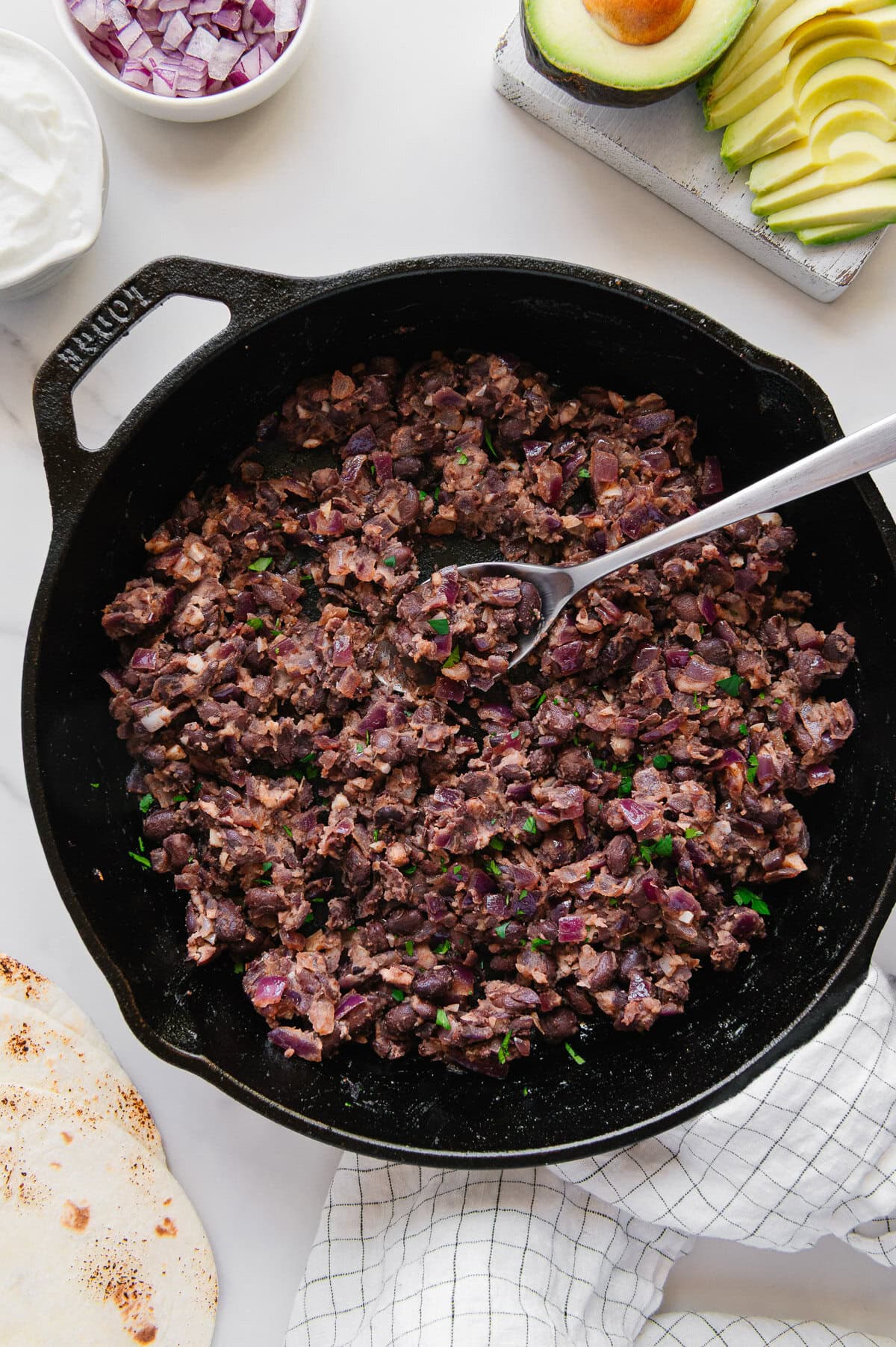 A cast iron skillet filled with cooked, mashed black beans and diced onions, with a spoon resting inside. Nearby are sliced avocado, diced red onion, sour cream, a tortilla, and a checkered towel.