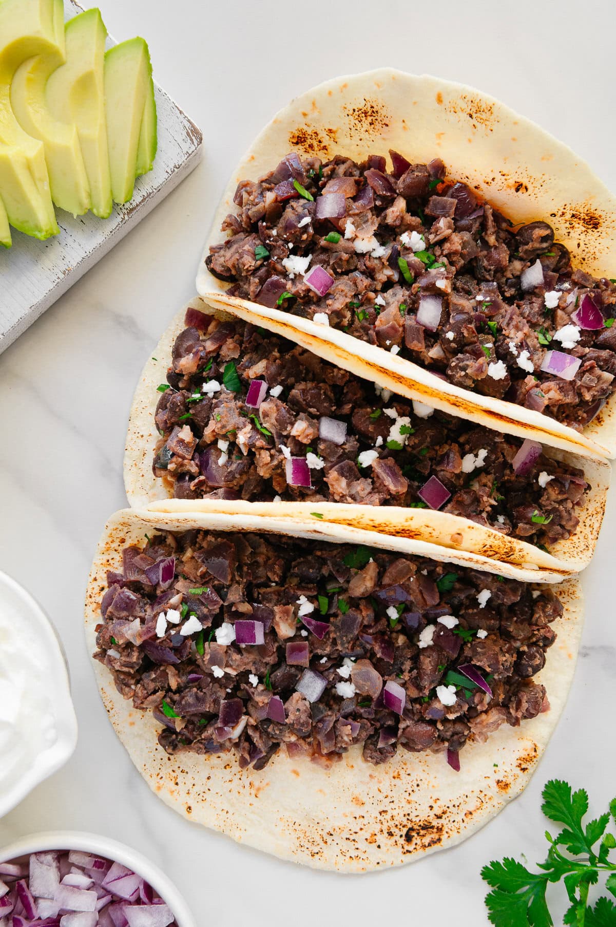 Three tortillas filled with refried black beans, chopped red onion, and crumbled cheese, displayed on a white surface. Sliced avocado, cilantro, and sour cream are arranged nearby as garnishes.
